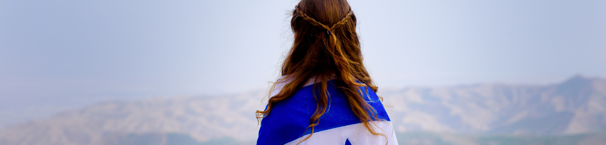 Young woman with brown hair atop a mountain looking at the view, wearing the Israeli flag as a cape.